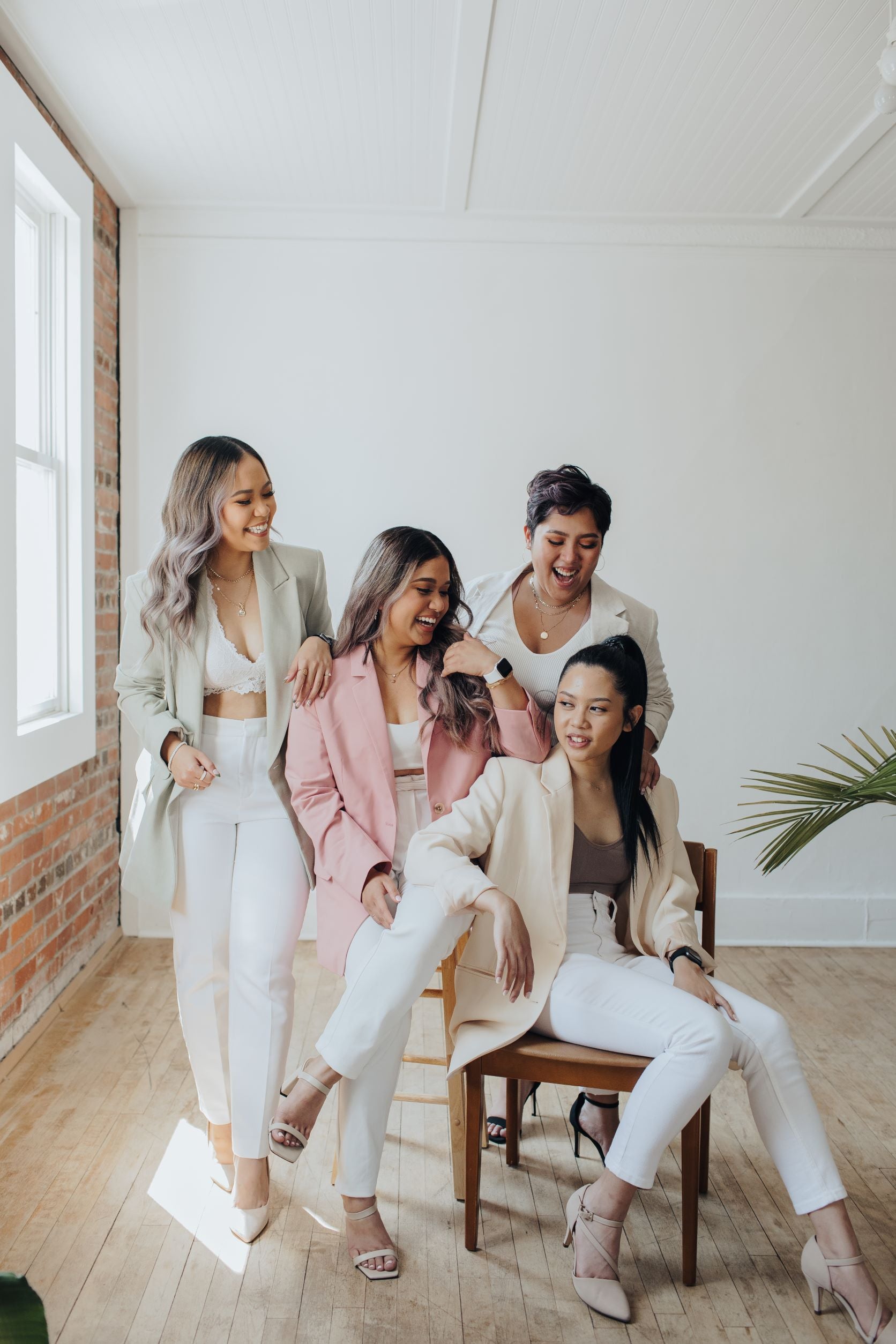 one lady sitting on a chair on the right, with another lady behind her and another girl in the middle leaning on the lady sitting on the chair and a lady standing on the left leaning on the lady in the middle